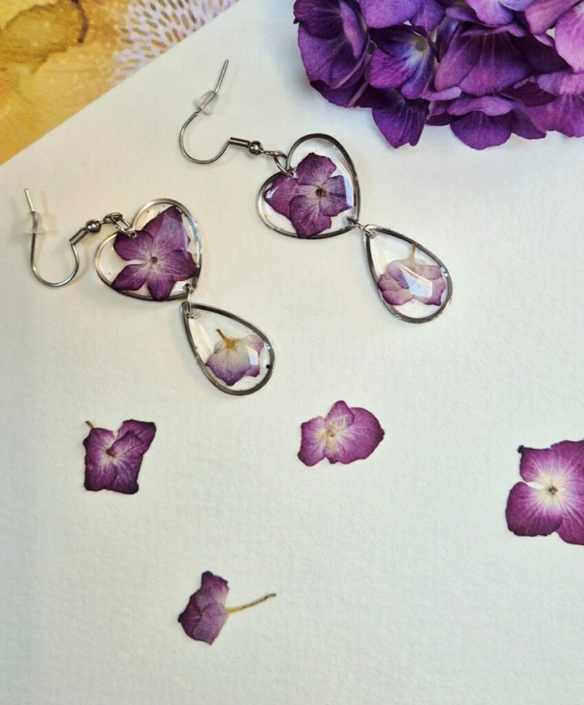 Boucles d'oreilles cœur et goutte en résine contenant des fleurs violettes séchées, entourées de quelques fleurs posées sur le papier