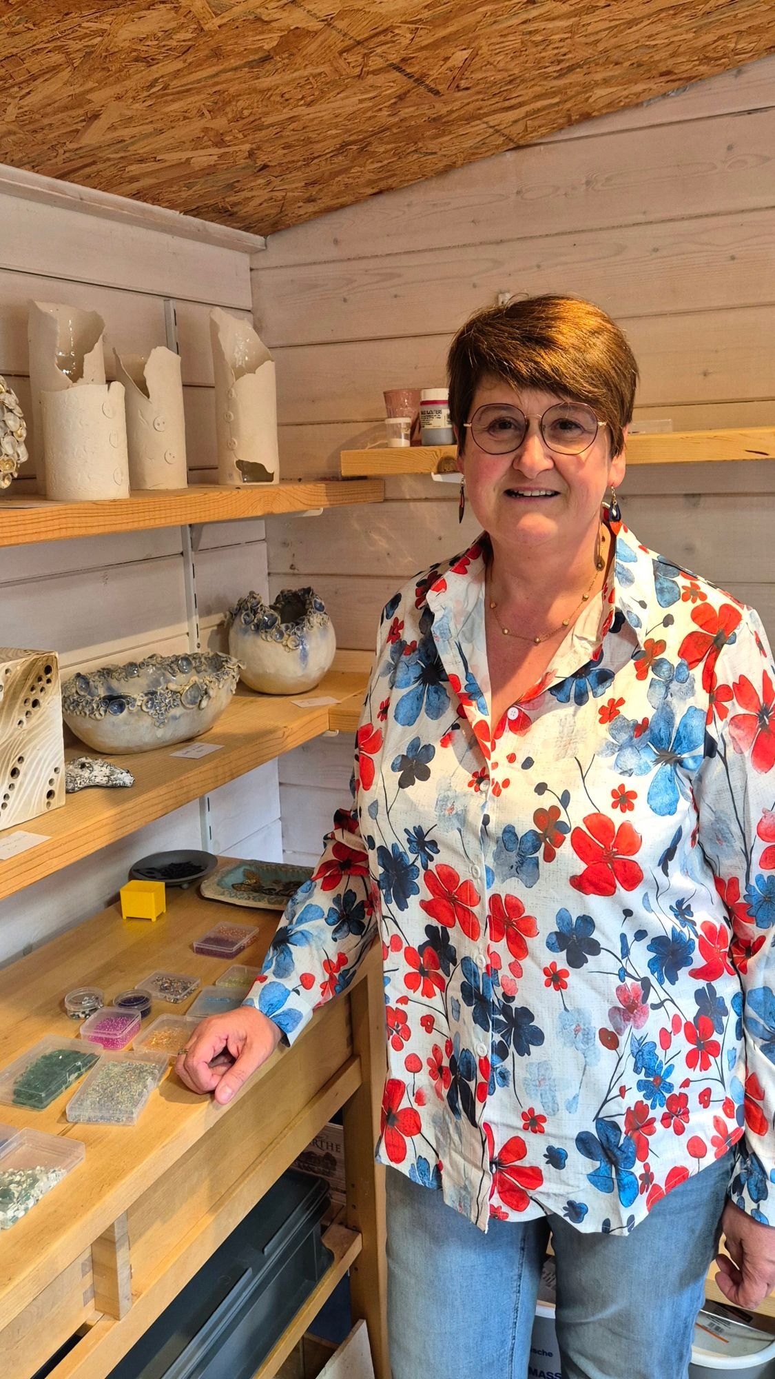 Chantal souriante dans son atelier de Beauvechain, devant des étagères de céramiques et ses boîtes de perles