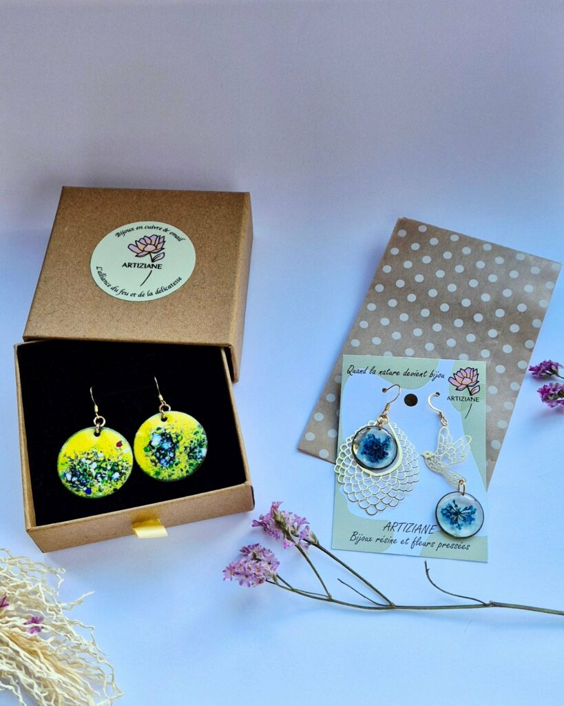 Boucles d'oreilles en émail jaune dans leur écrin Artiziane, et boucles en résine aux fleurs bleues sur leur carte, devant une pochette en papier