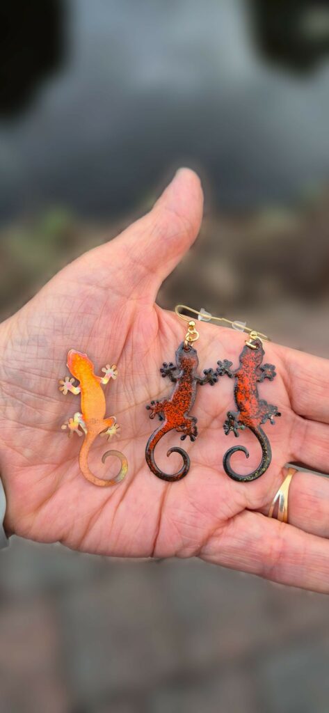 Dans une main, un gecko en cuivre brut à côté d'une paire de boucles d'oreilles gecko émaillées orange et noir