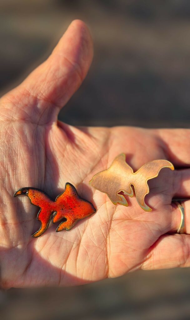 Dans une main, un poisson d'or en cuivre brut à côté d'un poisson d'or émaillé orange et rouge