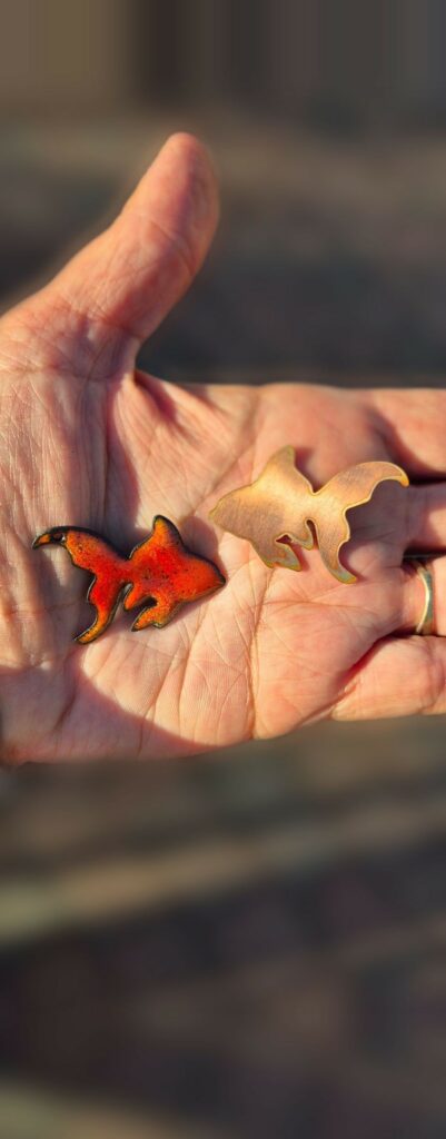Dans une main, un poisson d'or en cuivre brut à côté d'un poisson d'or émaillé orange et rouge